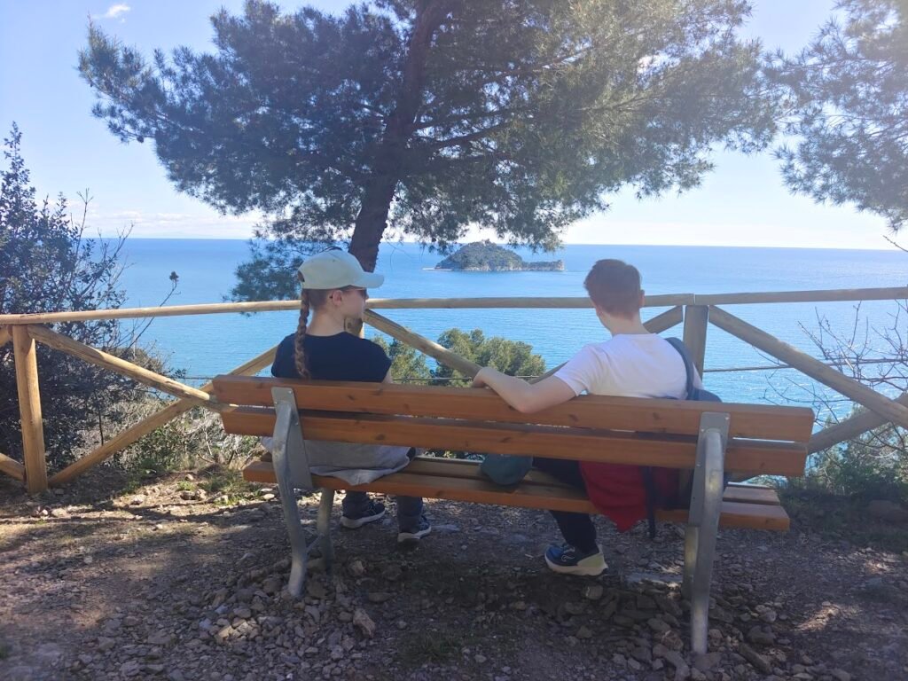 Due persone sedute su una panchina lungo la Via Julia Augusta ad Albenga, con vista sul mare.